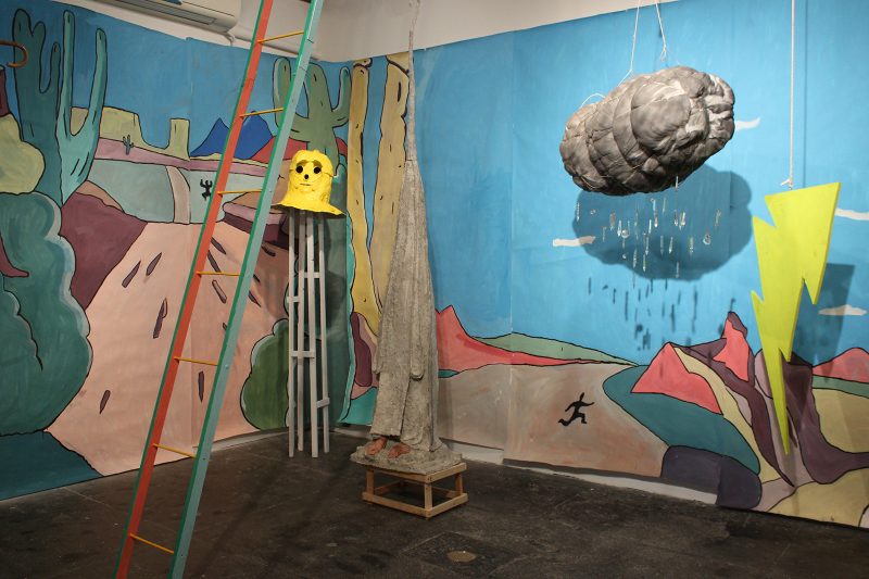 Installation view of Matt Freedman's "SLAP-STICK" including "Golem Helmet," desert panorama, ladder, dark cloud, and lightning bolt.
