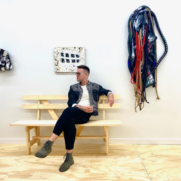 Alex Connor, founder and Director of Commonweal Gallery, sits in the gallery holding a drink in front of art at one of his openings.