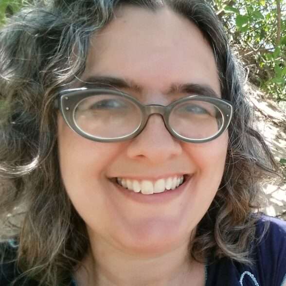 A close up of a woman's smiling face. She's wearing glasses and her curly hair frames her face.