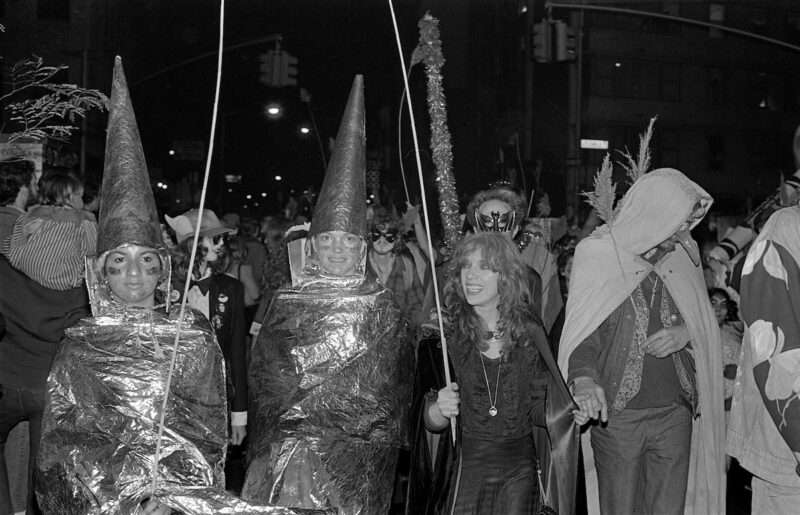 In a black-and-white photo, a group of fanciful, costume-clad Halloween celebrants parade down a New York City street. 