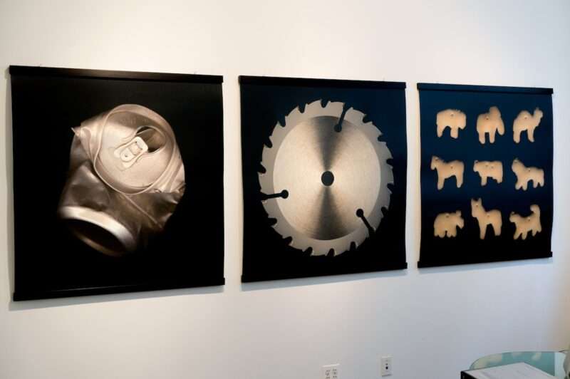 A white gallery wall shows three large photographic prints of objects placed in front of deep black backgrounds, from left to right is a crushed soda can, a saw blade and a grid of nine animal cracker cookies.