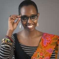 A Black woman with short cropped hair smiles widely and touches her black eyeglasses with her right hand like she’s adjusting them, she is wearing a black and grey striped shirt, colorful bracelets and scarf and prominent white earrings.