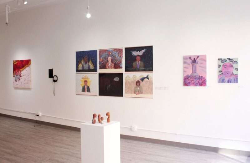 A white-walled gallery is installed with paintings on the walls, small sculptures on a pedestal on the floor in front of the paintings and a small video with earphones also on the wall. 