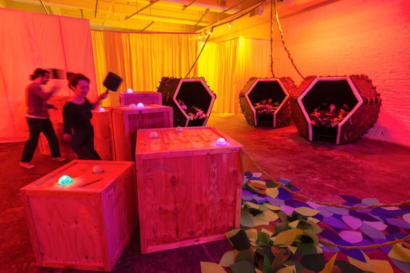 In a large space washed in a pink, yellow and red glow, three people rest in three comfort pods on the right, and two people wield mallets and try to hit lighted colored balls in wood chests on the left. This is an art game by Risa Puno that suggests all of us are care givers and care receivers.
