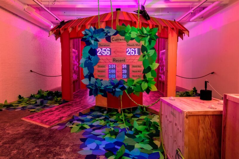In a large space washed in a pink, yellow and red glow, is an installation of a game representing care-giving, with a native hut with a scoreboard showing numbers of “Needs Met” and times (“Duration”), indicating how many caring acts have been delivered and in what time frame.
