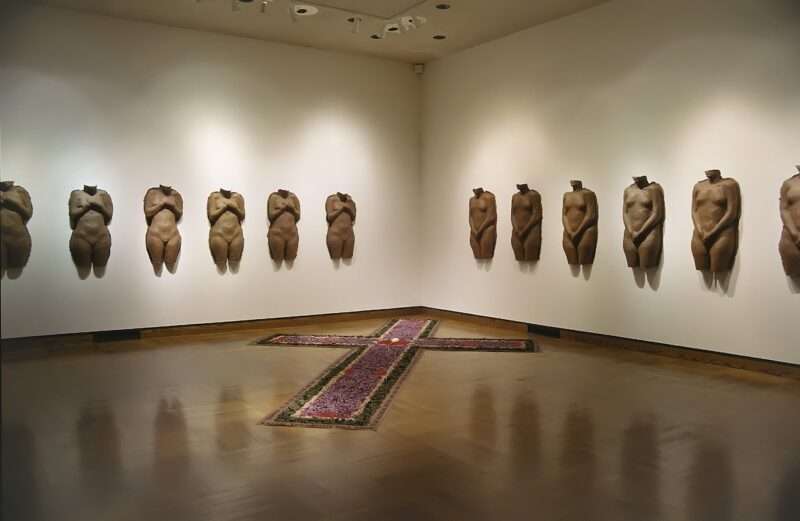 The corner of a gallery shows 12 exact same cast paper sculptures on two adjoining walls of a female torso, on the left wall hands covering her breasts, on the right wall, hands covering her vaginal region and on the polished wood floor, a sculpture of a Christian cross made from mixed media