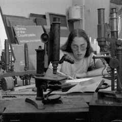 A black-and-white photo shows a young white woman artist with long hair parted in the center and wearing eye glasses working on a table filled with antique microscopes, concentrating intently on drawing one of them, a pencil in her right hand.