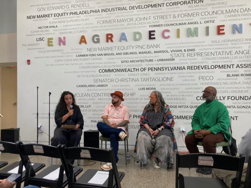 Four people sit on chairs in front of a wall filled with names of donors and others who have helped the event’s host, Taller Puertorriqueño. 