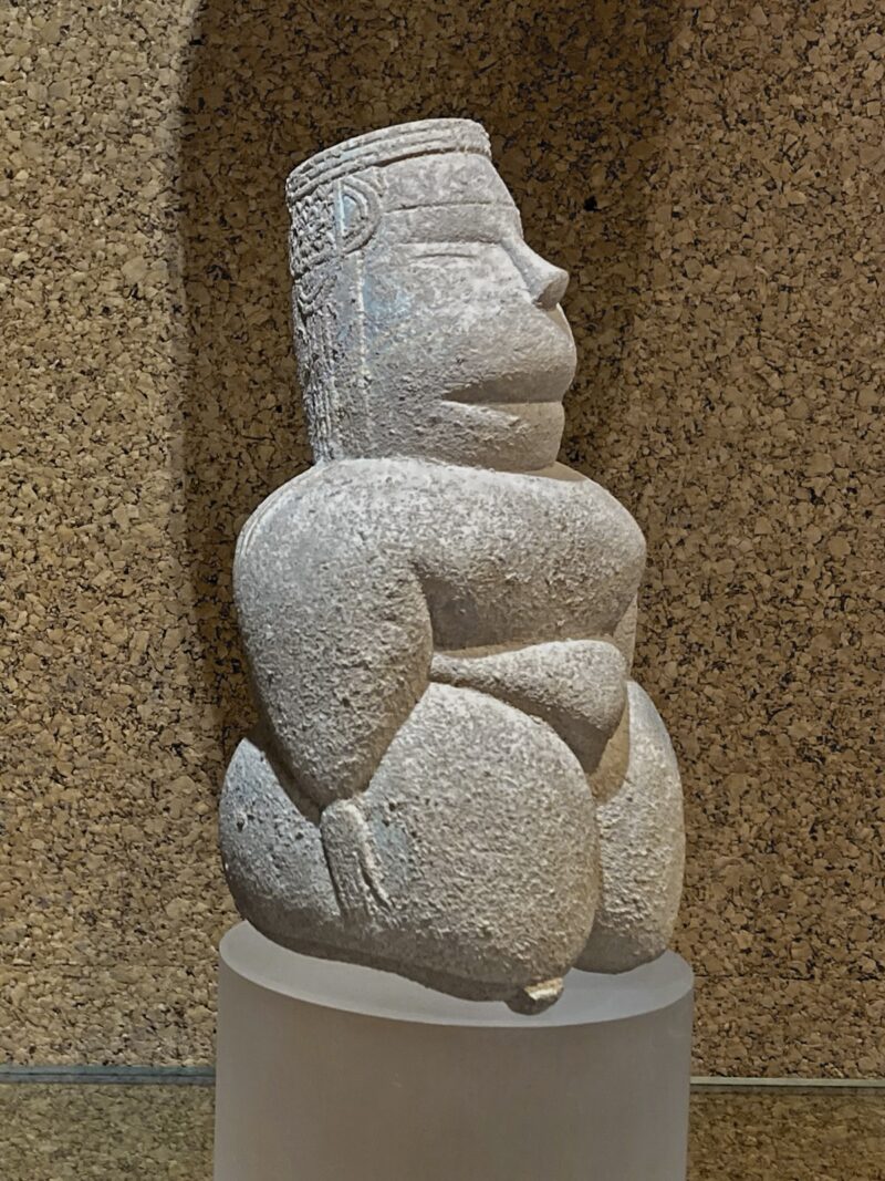 A carved stone statue from the Stone Age civilization on the island of Sardinia shows a woman, a goddess, who is kneeling, eyes closed, arms at her sides and wearing a patterned crown on her head.