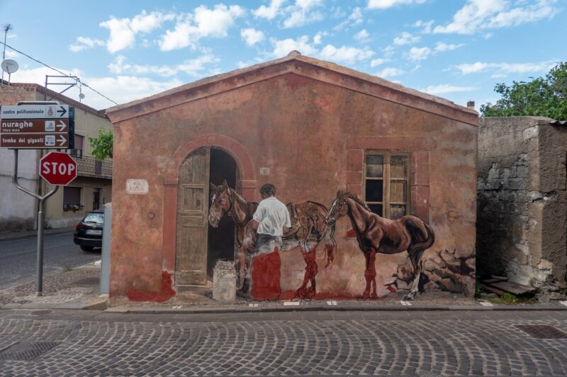 A crossroads of a town, Tinnura, Sardinia, with roads of cobblestone, shows a pitched roof one story building with a trompe-l’oeil mural painted on it of two horses and a man who is tending them in front of a partly opened wood door. 