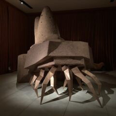 A hermit crab-like sculpture with long spider-y legs in front and a stepped series of shells on top suggests many legs carrying something very heavy and moving forward to a goal.