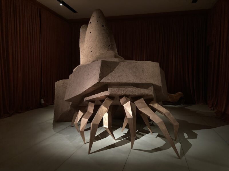 A hermit crab-like sculpture with long spider-y legs in front and a stepped series of shells on top suggests many legs carrying something very heavy and moving forward to a goal.