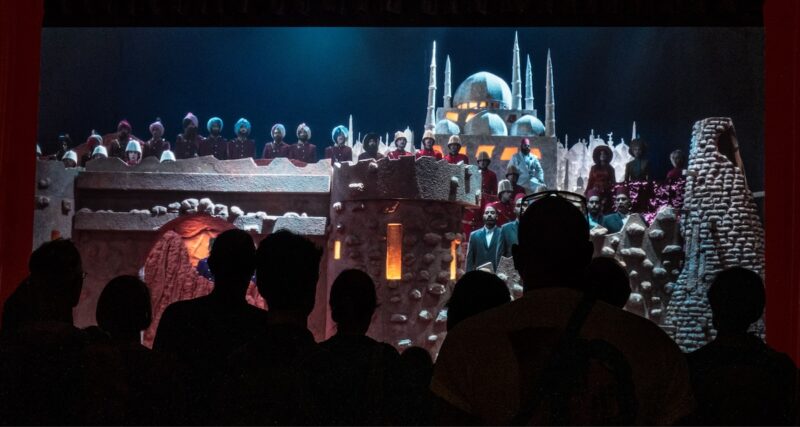 A stage set for an opera in progress, showing a night time scene of an ancient castle with turrets, and colonial soldiers standing on the roof. An audience of dark silhouettes stands in front.