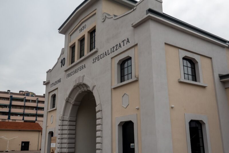 An old building with an arched doorway and square windows, whose facade looks like grey stucco with cream-colored touches, has Italian words near the top, “Station, Fricorifera, Specializzata.”