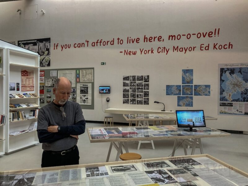 In a large high-ceilinged room in an art museum, a man wearing a grey t-shirt over a black, long sleeve shirt and black pants stands looking down at materials displayed on a table. 