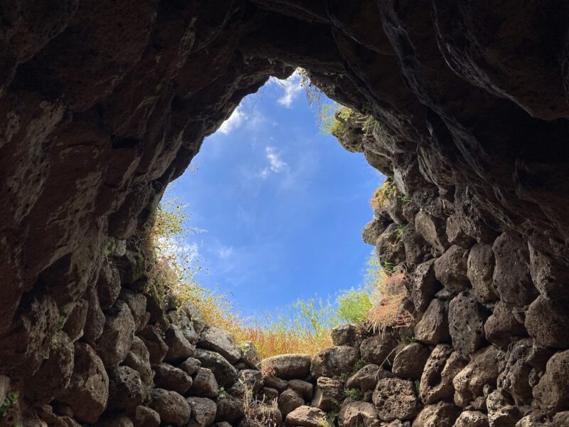 A cave-like structure with a round opening that shows the sky and some grass outside was made by Bronze Age humans who piled up accumulated rocks to make domiciles and religious structures, on the island of Sardinia.