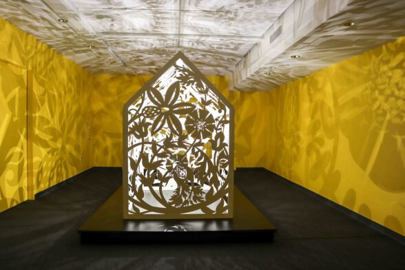 A 3-D outline of an archetypical house with open sides heavily embellished with floral cutouts, and empty inside but for bright lights that cast ghost images of the floral patterns on the three yellow-painted walls and white ceiling.