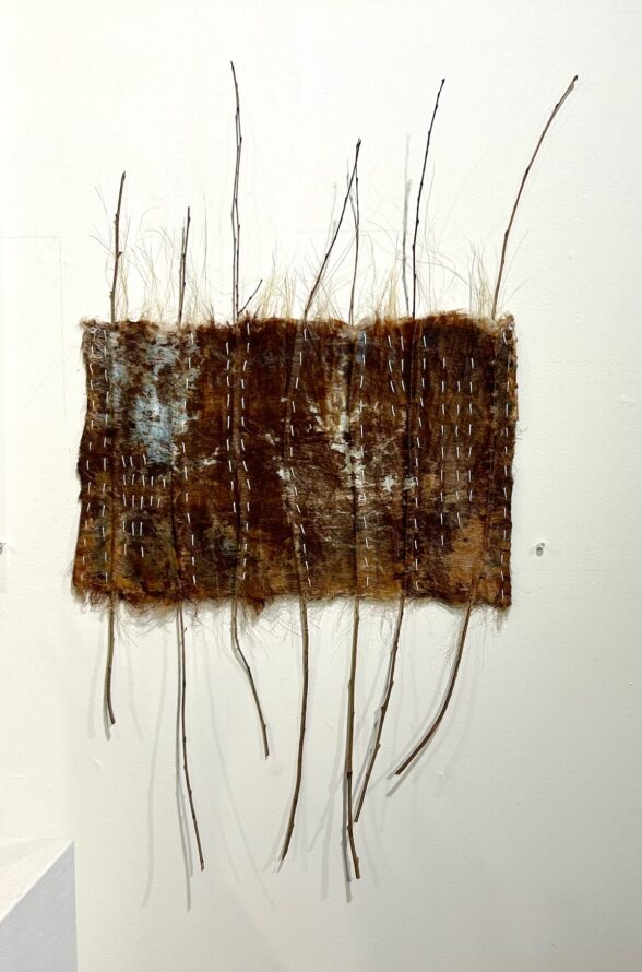 A dark, animal-pelt-like fiber art work sits on a white wall, with thin twigs running through the fiber and sticking out on top and bottom and with hand stitching in white thread that funs vertically mimicking the twigs movement through the piece. 