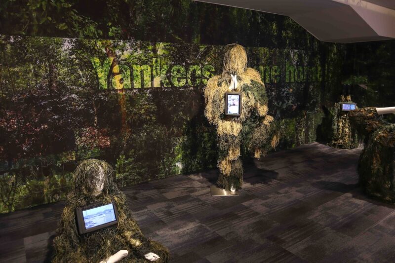 In a dark space, a dramatic scene shows camouflaged store mannikins in front of a blown up photo of a deep jungle scene on the wall, each figure in a yoga pose with small video monitor showing images appended to them while also on the wall are letters superimposed on the jungle photo saying, “Yankees come home.”