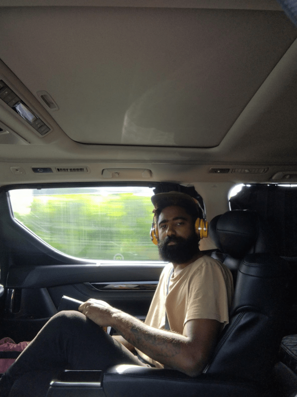A Black man with a beard wears orange ear phones and sits as a passenger in a fast-moving vehicle that looks like a van with black comfy seats.