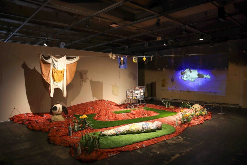 An installation in a museum shows red and green fabric sleeping bags on the floor, laundry hung on a line above, a shopping cart typically used by the homeless, what looks like a dead body wrapped in cloth and paper in the foreground and a video projection on the wall.