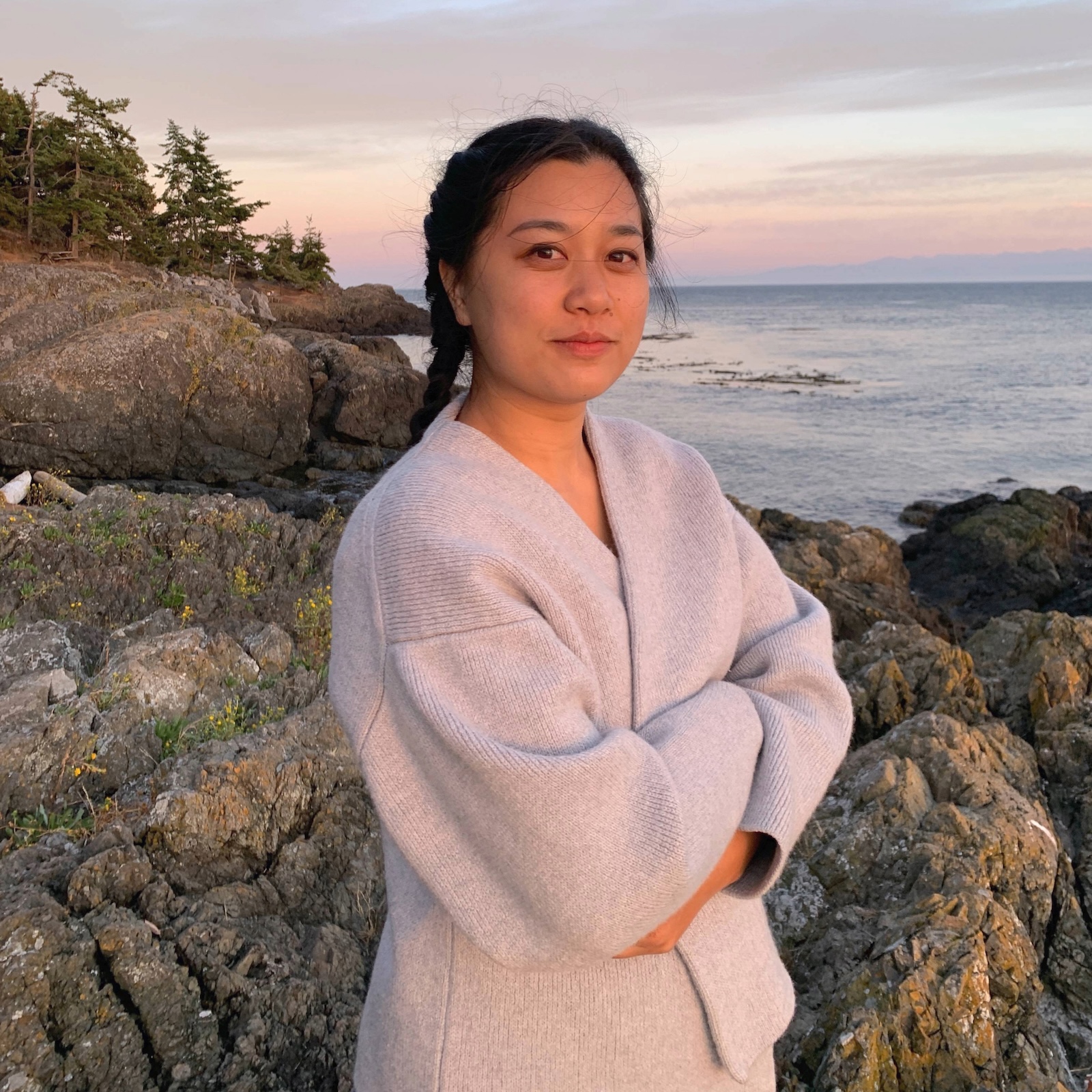 A young Asian woman stands in front of a rocky shoreline with pine trees in the left background and the ocean in the right, and she smiles at the camera and looks content, her arms crossed over her chest, and her body covered in a comfy grey sweater.