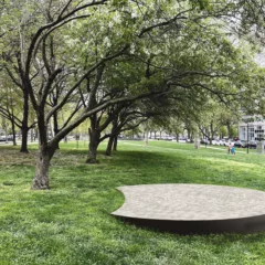 A park shows a grove of trees in a grassy space in the middle of several streets with cars parked and driving. In the foreground is a low platform of river stones embedded in concrete in the form of a kind of circle with a “smile” cut out of one side.