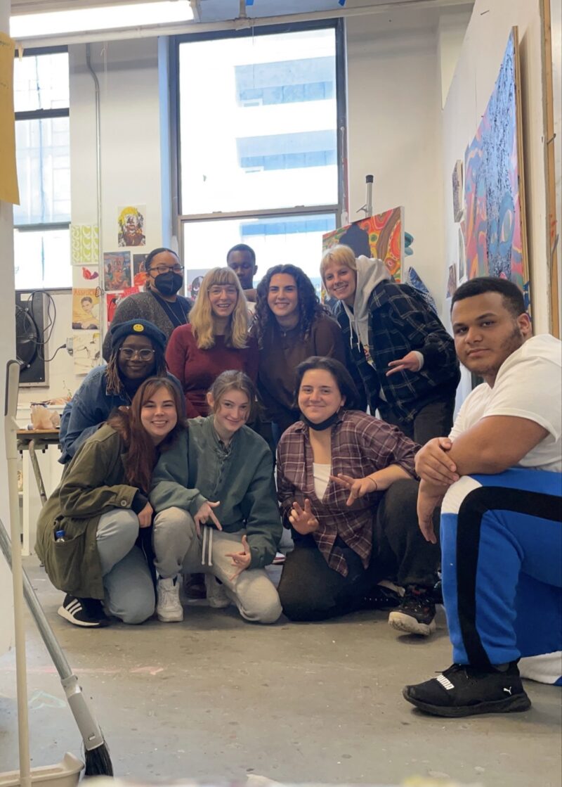 A group of art students poses, crouched on the floor, for a photo in a painting studio, in 2023, and they all look happy, smiling and relaxed. The students were juniors, painting majors.