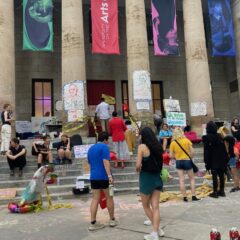 People on steps and the sidewalk in front of a building with large pillars and banners flying between them. They are students and their university has just announced it is closing.