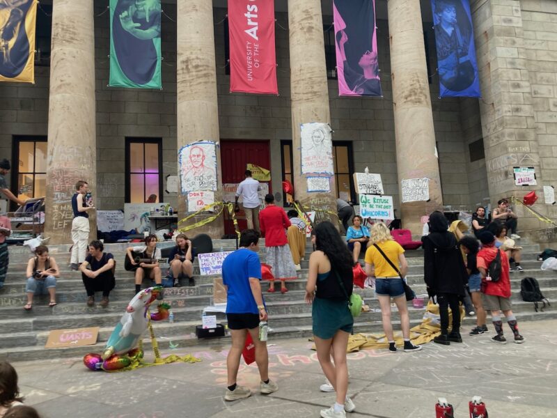 People on steps and the sidewalk in front of a building with large pillars and banners flying between them. They are students and their university has just announced it is closing.