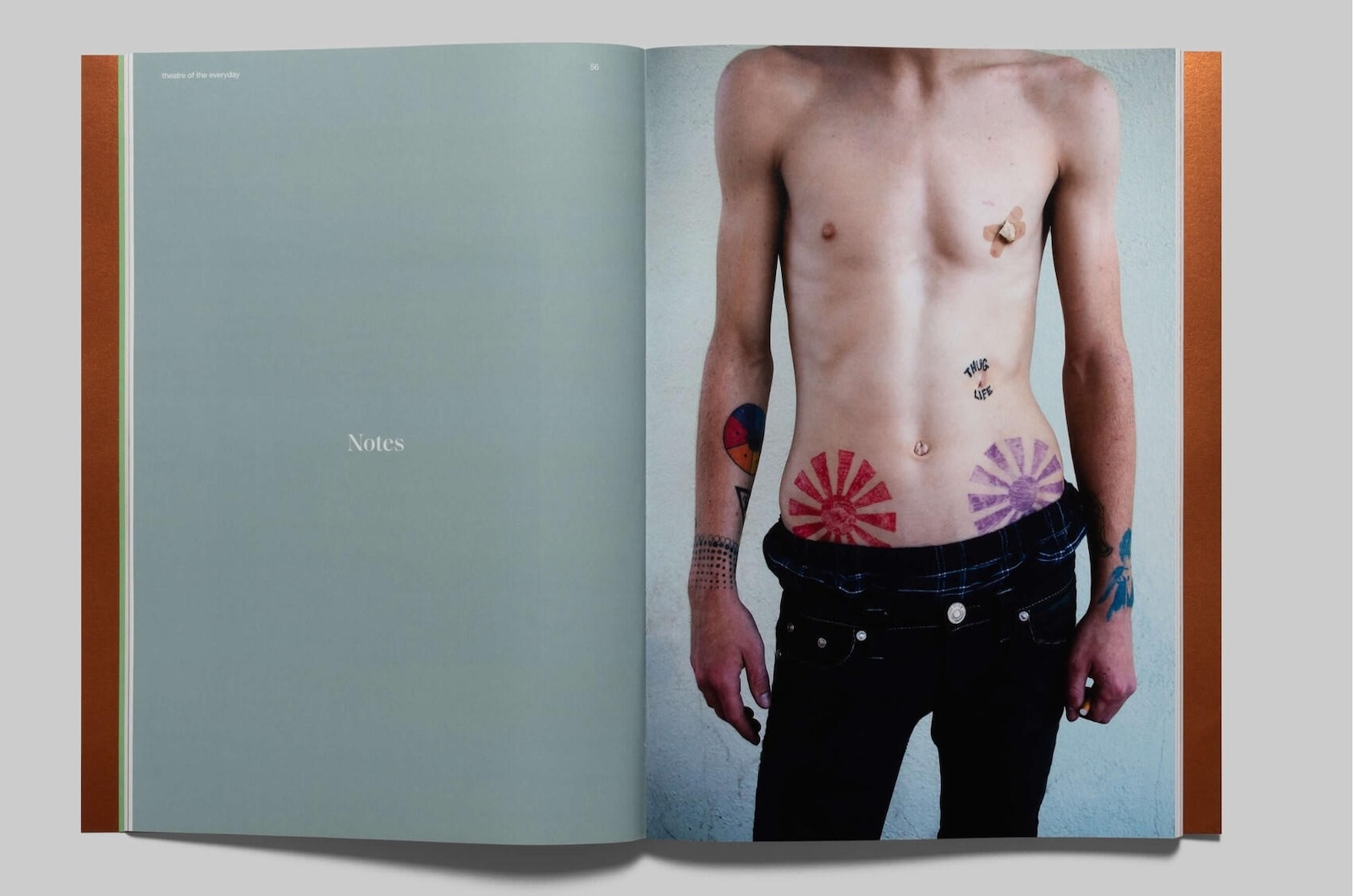 A two-page spread in a book shows the left hand page all grey with the word “Notes” in white, centered on the page, and on the right-hand page, a man’s nude torso from collarbone to hips shows a tattooed body with two small bandages crossed over the man’s left nipple; his black pants set off the pale skin.