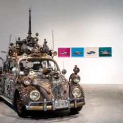 In an art gallery with a concrete floor and white walls sits an ancient VW bug car with Maryland plates and all manner of adornment welded to the exterior, including lights, beads, chains, decorative spires and egg shapes.