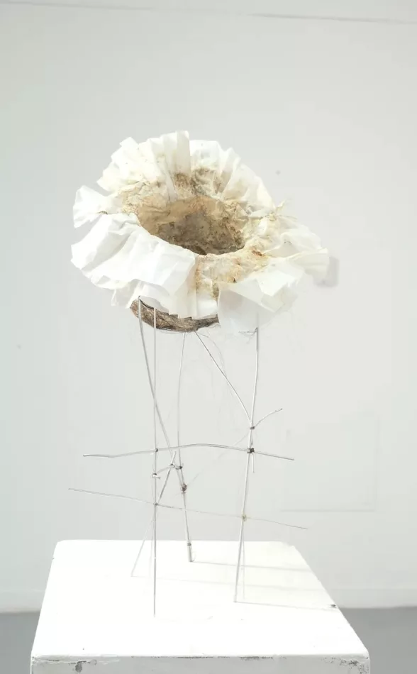 A delicate-looking flower-like object with all white petals that surround a large dirty hole in the middle sits precariously on a group of gerry rigged thin aluminum rods, making it look unstable and ready to fall over.