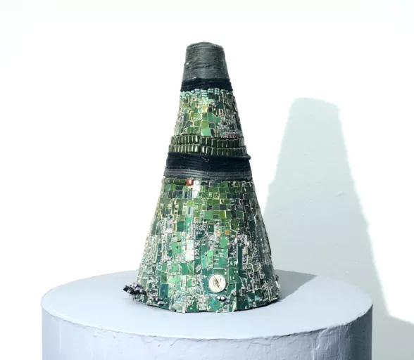 A cone-shaped object made from cut up circuitry and other material sits on a pedestal, looking like a small monument. A traffic cone is suggested but the circuitry chips give the piece a festive air.