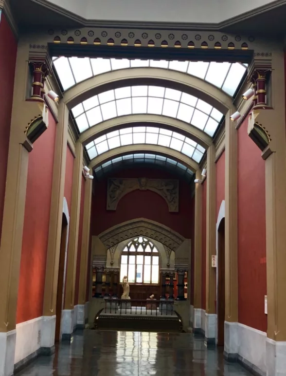 A photo shows an historic art museum with a arched skylight in a hallway bordered by walls painted a warm rosy red, and in the distance an arched window sets in a wall behind several statues and other art. The shot of of the interior of PAFA.