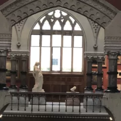 A photo shows an ornate interior of an art museum with arched leaded glass window and chiseled stone framework over the window, and in the foreground a white marble statue of a female in a classical robe and with casually twisted posture.