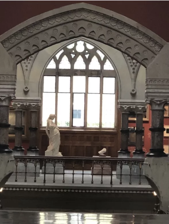 A photo shows an ornate interior of an art museum with arched leaded glass window and chiseled stone framework over the window, and in the foreground a white marble statue of a female in a classical robe and with casually twisted posture.