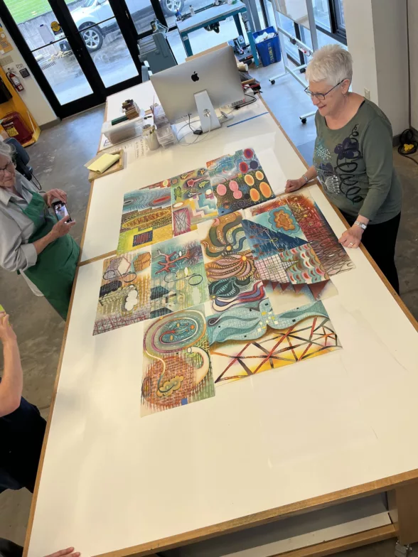 A workshop room shown in birds eye view, has a grey-haired woman on the right talking about her printed art displayed on a long white table and on the left are several students who are looking at the works.