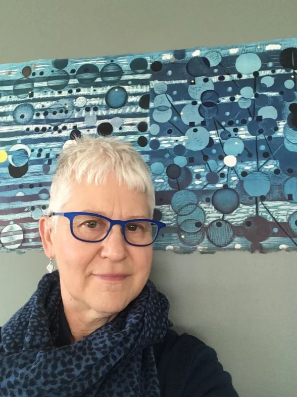 A smiling woman with short white hair, stylish blue-rimmed glasses and a stylish blue dotted neck scarf stands in front of a wall, with lines and dots in shades of blue and black which echoes the woman’s accessories. 