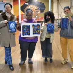 In an indoor space with wood floors and a mural in the background, four people dressed in coats and long pants stand facing the camera. Between their hands, they each hold a dark blue cyanotype print with white figure drawings and colorful marks.