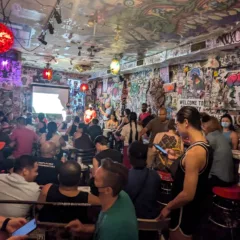 Shown from the back of the space, a full room of people are engaged in conversation with their tablemates, as three presenters stand in the front. The walls of the room are covered with tags, signs, and stickers, and colored lighting fixtures dot the space.