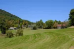 A landscape shows a large undulating field of grass dotted with trees and in the background on the left a tree-covered hill and at the base of the hill several large buildings.