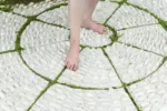 Two bare feet and legs are shown below the knees, with the person standing on a white round rug-type sculptural object inset into the grass in a park.