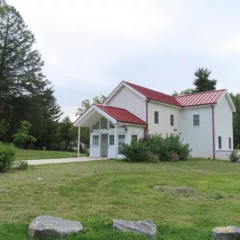 A renovated historic farm house looks inviting, with a brand new red roof and gutters and many windows and a nice vestibule entryway led up to with a concrete walkway bisecting grassy spaces and trees to either side.