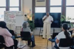 A group sits in a seminar room with large windows, and at the front is a Black, non-binary speaker wearing black pants and top, who is leading the seminar, speaking into a mic.