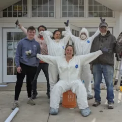 A joyous group of volunteers, some in white hazmat suits, congregate for a jubilant photo in front of an historic farmhouse that they helped renovate into an art space for the community.