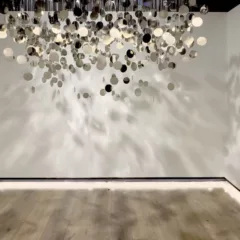 An art installation of mirror-like metal discs is hung by filaments from the ceiling above and light cast on the piece create shadows on the three surrounding walls and floor.