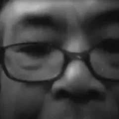 A fuzzy black and white photo shows an extreme closeup of the eyes, eyebrows, dark eyeglasses and nose of a person surrounded by a dark background but with some light spots on the nose and forehead.