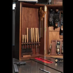 A close up of some woodworking tools in a nice wood frame on top of a metal table with more tools on it. dark room with a few bright spots shows a stack of skateboard decks behind a partial wood wall with a blueprint of a building tacked to it. The space is mysterious and looks unfinished and is part of a larger installation.