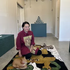 An interior art space with concrete floor, white walls and a a glass-paneled garage door in the background shows a woman with a long dark ponytail sitting cross-legged on a cat-patterned rug surrounded by three green benches.